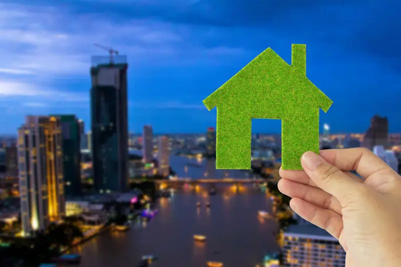 Symbol für nachhaltiges Bauen: Eine Hand hält ein grünes Haus-Icon vor einer modernen Stadtlandschaft bei Nacht. Das Bild steht für umweltfreundliche Bauweise und Energieeffizienz.